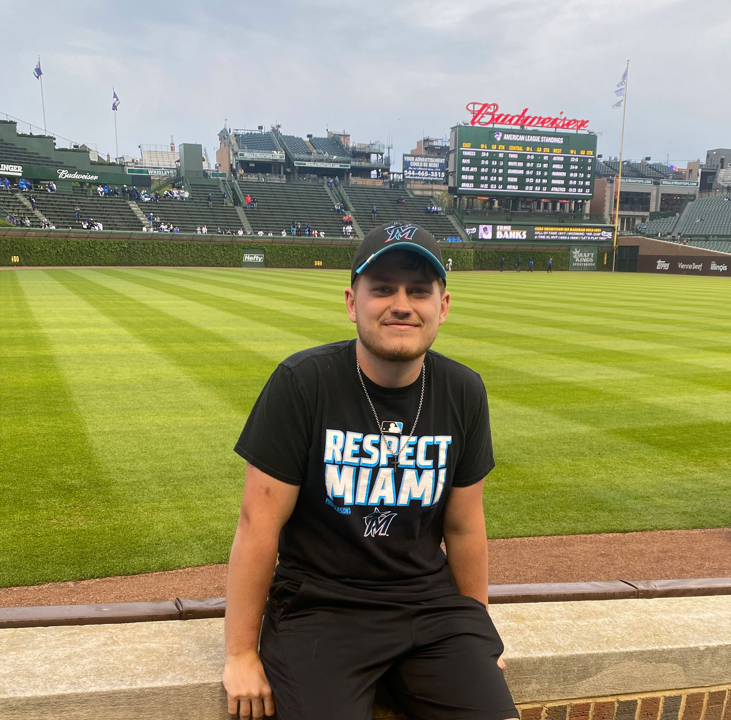 A man wearing a baseball cap, black shirt and shorts sitting on a ledge with a baseball stadium behind him.