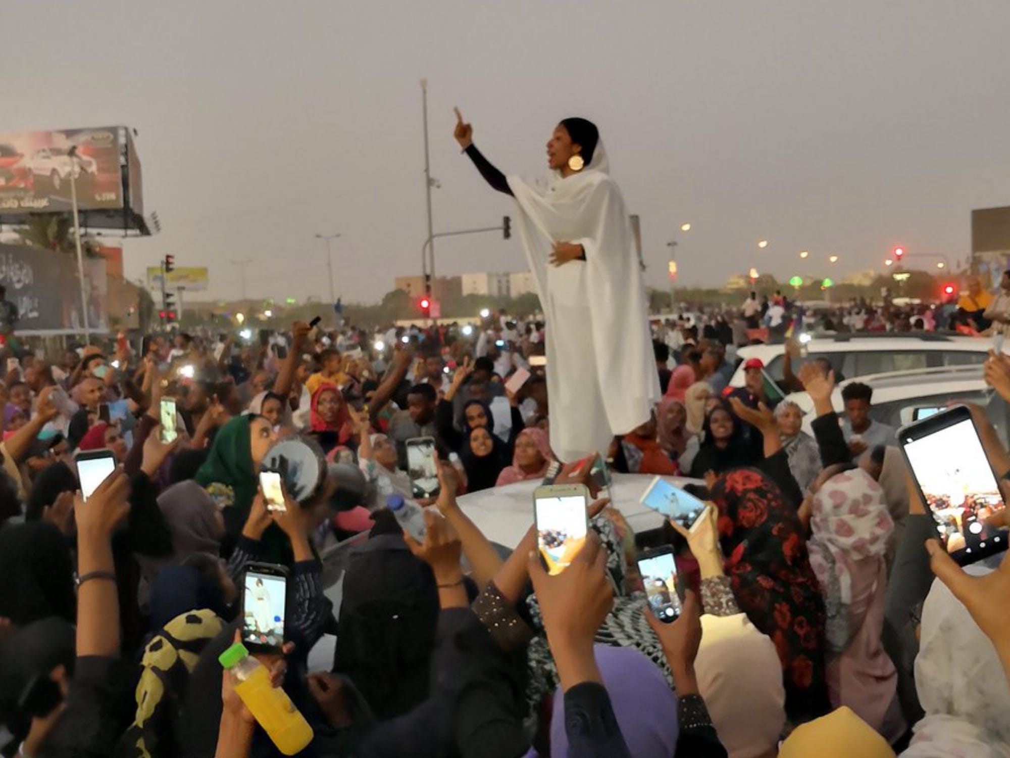 a crowd of cell phones raised to a woman, clad in a white hijab, falling back as she speaks to a crowd.