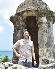 man wearing a tan shirt smiling at the camera in front of an old ruin of a tower