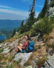 A woman in hiking gear smiling at the camera as she sits on the side of a mountain