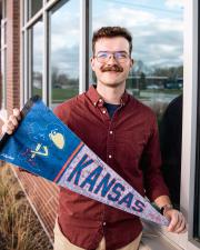 A man with a red shirt and glasses holding a University of Kansas Pennant that shows the old 1912 Jayhawk illustration.