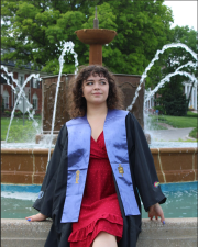 A woman wearing graduation attire, in front of a fountain, looking off into the distance