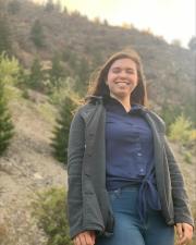 an angled shot of a woman wearing a jacket, smiling at the camera with rolling hills spotted with trees in the background