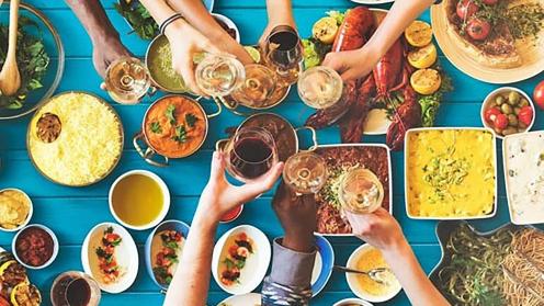 an aerial image of a bright blue table clad with colorful orange, yellow and purple dishes with a diverse group of arms cheers-ing wine glasses
