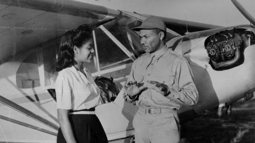 In front of an old airplane stands African American woman smiling at a African American man speaking. 