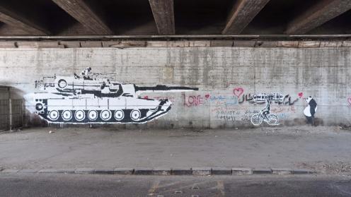 Graffiti under a bridge depicting a black and white graffiti tank on the move, pointed towards a black and white profile image of a child riding a bike and a panda.